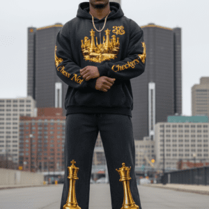 A man standing in downtown Detroit wearing the 313 Emblems "Chess Not Checkers" bundle. The black hoodie features a metallic gold chess piece crown graphic on the chest and "Chess Not Checkers" script in gold along the sleeves. The matching black joggers feature large metallic gold King and Queen chess piece graphics on the lower legs.