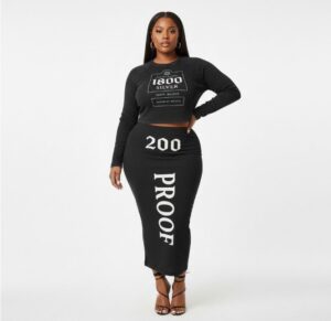 A studio shot of a woman wearing the 313 Emblems "1800 Proof" two-piece set. The black long-sleeve crop top features an "1800 Silver" tequila-inspired graphic, paired with a matching black midi skirt featuring "200 PROOF" printed in white Gothic-style lettering.