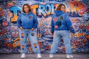 A woman wearing the 313 Emblems "313 Teams" royal blue hoodie and matching light-wash denim jeans. Both pieces are detailed with Detroit sports team patches and colorful vertical "DETROIT" branding, set against a graffiti-style city backdrop.
