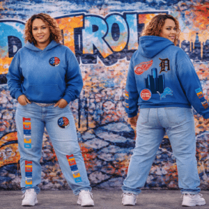 A woman wearing the 313 Emblems "313 Teams" royal blue hoodie and matching light-wash denim jeans. Both pieces are detailed with Detroit sports team patches and colorful vertical "DETROIT" branding, set against a graffiti-style city backdrop.
